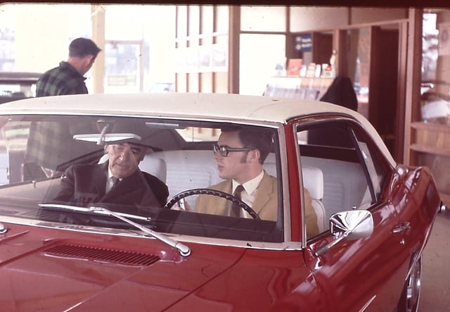 Two men sit in a red convertible in the showroom lot